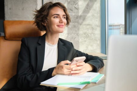 Portrait of young professional saleswoman, businesswoman in suit, sitting in office at her company, using smartphone, working on laptop